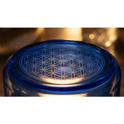 Ein blauer Glasbehälter mit einem aufwendigen „Blume des Lebens“-Muster, das in seine Oberfläche eingraviert ist, vor einem verschwommenen Hintergrund in warmen Farbtönen.