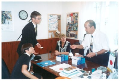 Eine Gruppe von Personen, vermutlich Lehrer oder Verwaltungsangestellte, hat sich um einen Schreibtisch versammelt, der sich offenbar in einem Büro oder Klassenzimmer befindet; im Hintergrund sind verschiedene Gegenstände und Dekorationen zu sehen.