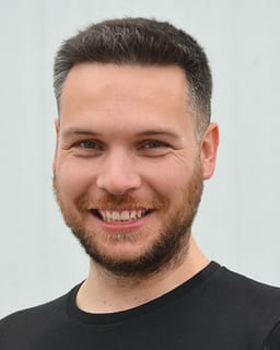 A smiling man with short brown hair and a beard is in the foreground, with a plain white wall in the background.