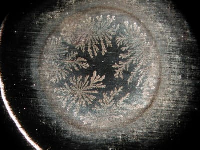 Des motifs cristallins complexes, ressemblant à des fougères, forment un agencement circulaire sur un fond sombre et texturé, délimité par un bord incurvé et lumineux.