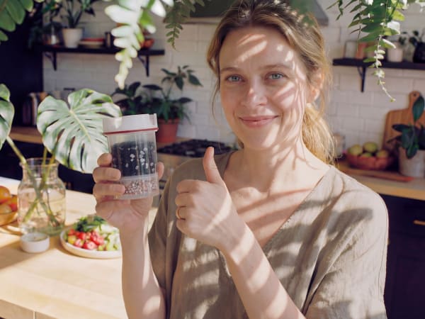 A young woman with blonde hair stands in a kitchen surrounded by various plants and fruits, holding up a glass jar.