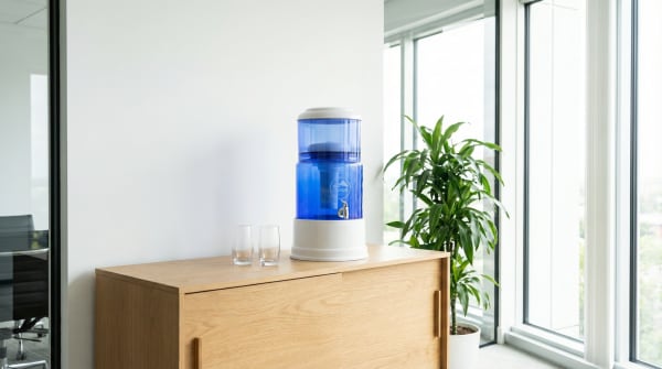 A blue water filter dispenser sits on a wooden cabinet, with a potted plant and large windows in the background, creating a clean and minimalist interior setting.