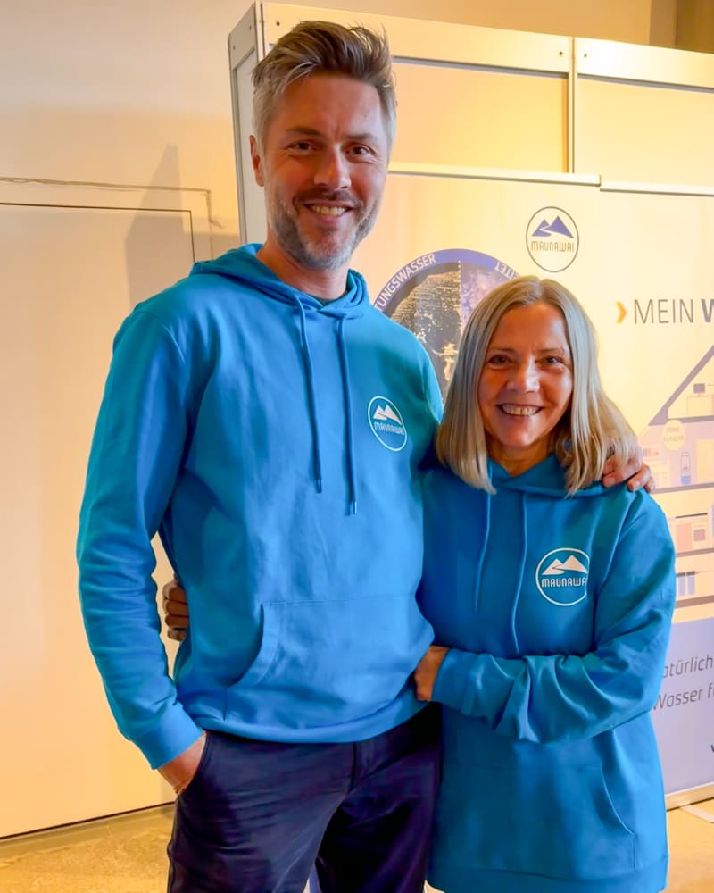 Un homme et une femme, vêtus de sweats à capuche bleus assortis, se tiennent devant un mur sur lequel figurent un logo et un texte.